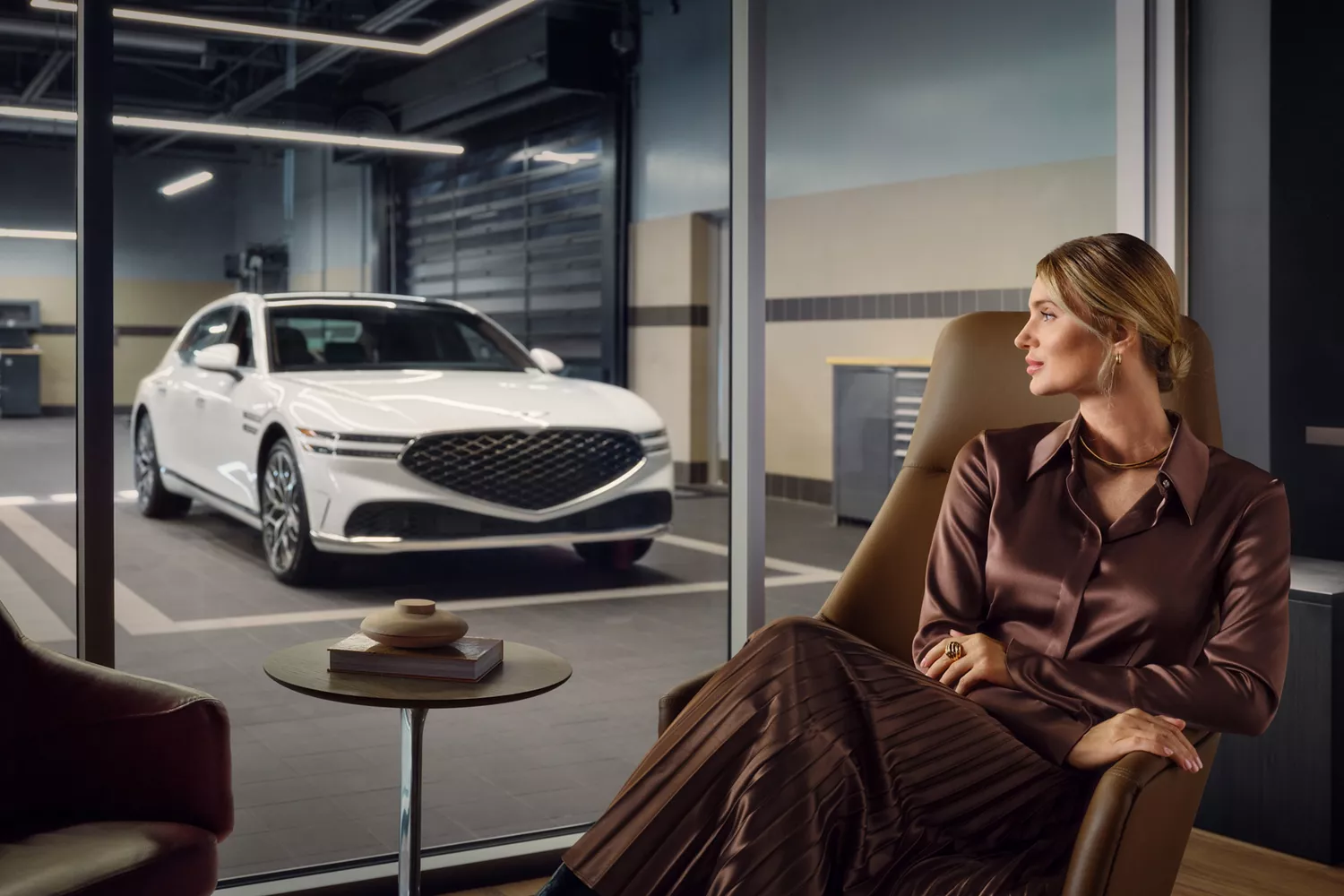 A woman in a brown outfit sits in a chair, looking at a white car parked in a garage through a window.